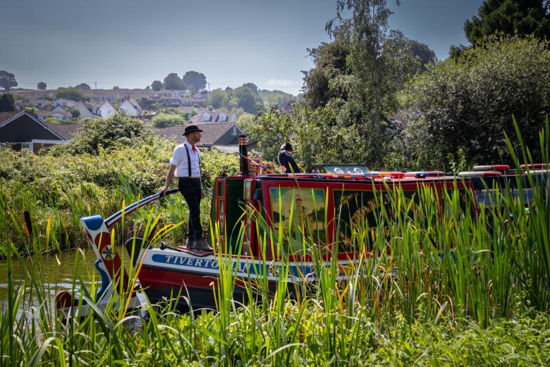 The canal boat being steered down the Great Western Canal at Tiverton. The man holding the rudder is in old English clothing.